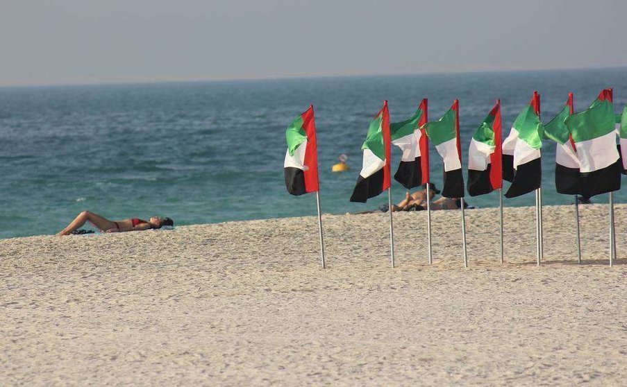 Bañistas toman el sol en la playa de Kite Beach en Dubai junto a banderas de Emiratos Árabes que ondean al viendo. (R. Pérez / EL CORREO)